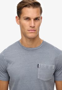 Grijze katoenen t-shirt met een ronde halslijn en korte mouwen. Heeft een linkerkant borstzak en een subtiel, gecapitonneerd stofpatroon.