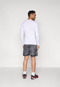 Λευκή αθλητική μπλούζα με μακριά μανίκια, συνδυασμένη με μαύρο σορτς που έχει υφή γκρι μοτίβου και μαύρο περίγραμμα. Φορεμένη με μαύρα και κόκκινα αθλητικά παπούτσια.