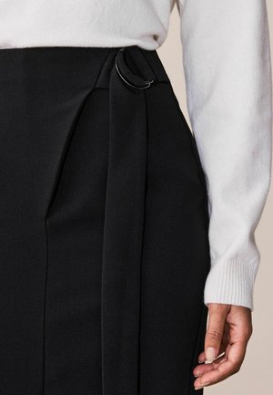 Zbliżenie czarnej spódnicy z bocznym paskiem i srebrną klamrą, zestawionej z białym swetrem z długim rękawem i swobodnie opadającą ręką po boku.