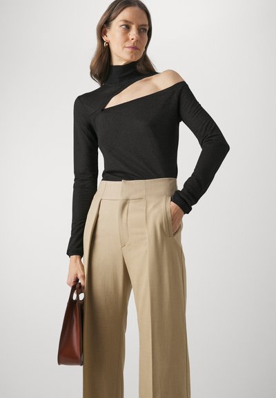 Zwarte langemouwen top met een asymmetrische uitsnijding, gecombineerd met hooggesneden beige broeken. Model houdt een bruine handtas vast.
