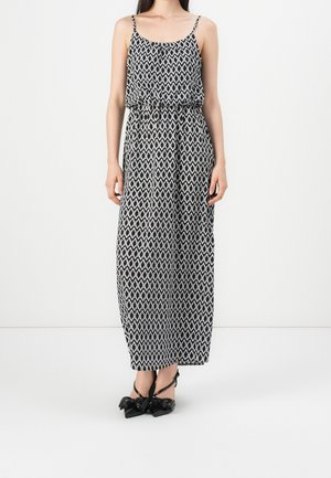 Femme portant une robe maxi sans manches à motif géométrique noir et blanc avec de fines bretelles et des talons noirs pointus avec des nœuds.