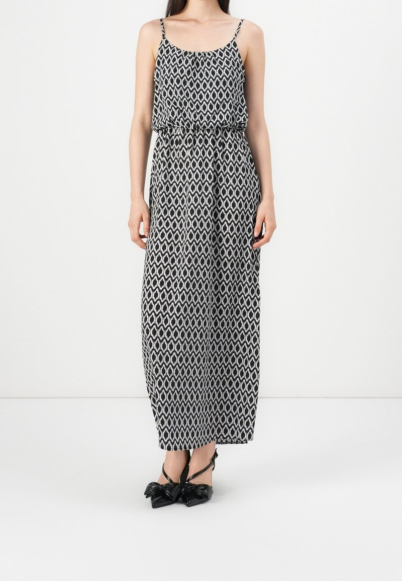 Femme portant une robe maxi sans manches à motif géométrique noir et blanc avec de fines bretelles et des talons noirs pointus avec des nœuds.