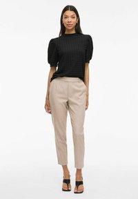Top nero strutturato con maniche a palloncino, abbinato a pantaloni beige su misura e sandali neri con design aperto.