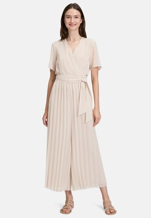 Combinaison plissée à effet portefeuille en tissu beige doux avec manches courtes et ceinture nouée à la taille, présentant des jambes larges et courtes ainsi qu'une texture lisse.