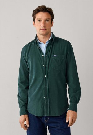 Hombre de pie con una camisa verde oscuro abotonada sobre una camisa azul clara con cuello y vaqueros azul oscuro sobre un fondo liso.