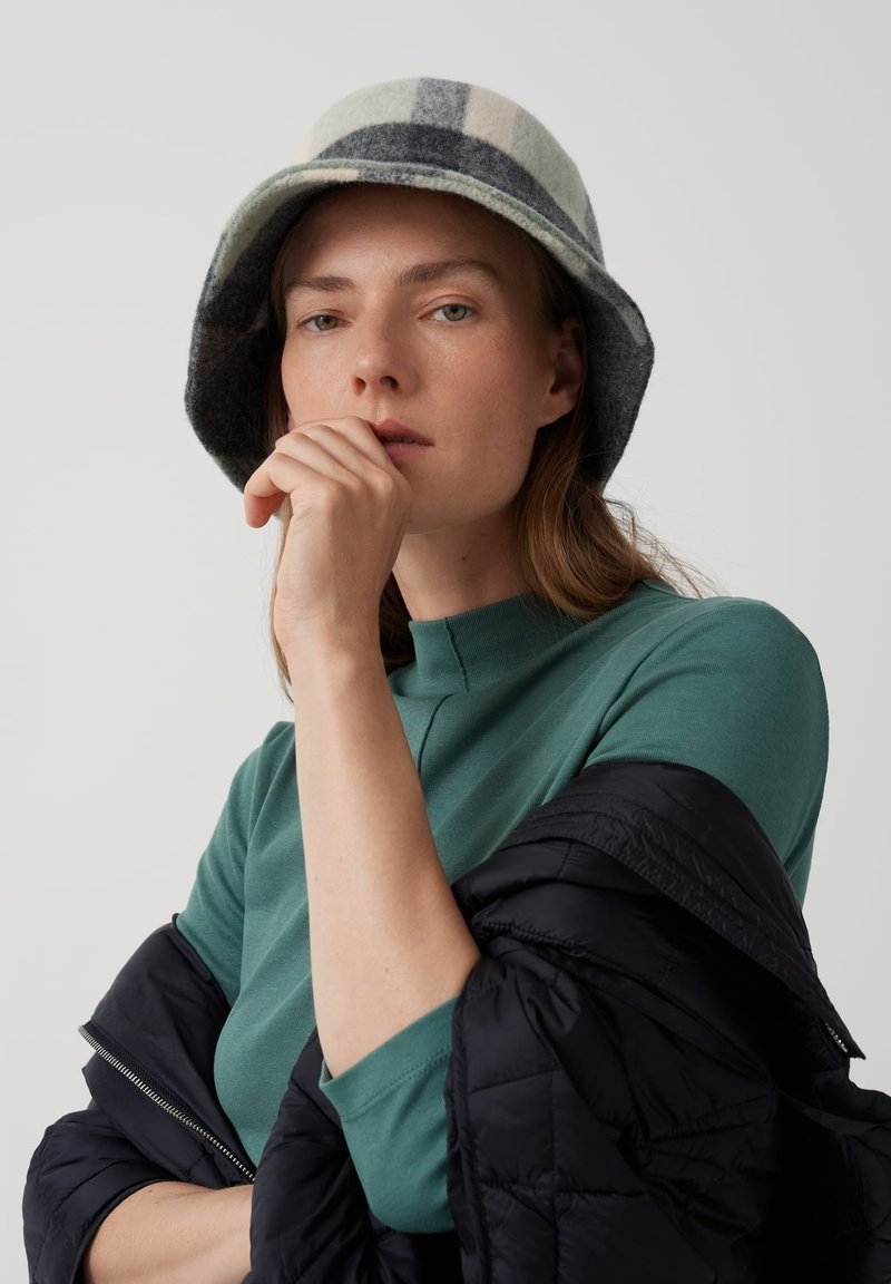 Wełniany kapelusz typu bucket w szarym kolorze z wzorem w kratkę; modelka ma na sobie turkusową bluzkę i czarną kurtkę puchową, trzymając myśląco podbródek.