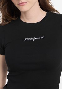 Crna rebrasta majica s prilagođenim dizajnom, s bijelim izvezenim logotipom "guessjeans" na području prsnog koša. Kratki rukavi i okrugli izrez.