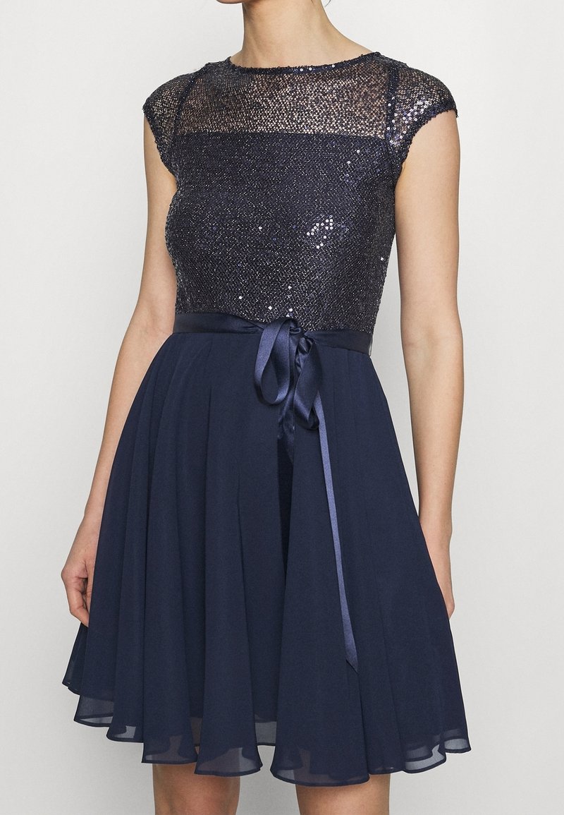 Robe bleu marine avec corsage à sequins, empiècement transparent, manches courtes et ruban en satin noué à la taille sur une jupe plissée en chiffon.