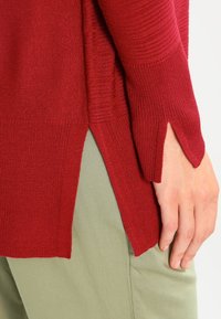 Czerwony dzianinowy sweter z bocznymi rozcięciami i teksturowanymi rękawami, zestawiony z jasnobeżowymi spodniami, ukazujący gładkie, miękkie wykończenie tkaniny.