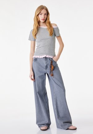 Jeune femme portant un haut gris épaules dénudées et un jean bleu à jambes larges avec un porte-clés tressé, debout, une main dans la poche.