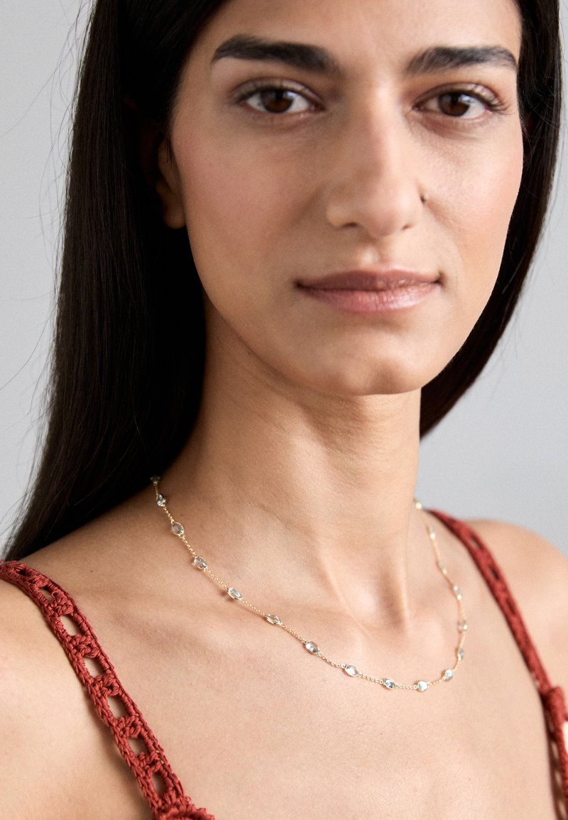 Zlatý náhrdelník se oválnými, průhlednými korálky, nošený podél klíční kosti, doplněný o červený top bez rukávů s háčkovanými detaily.