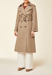 Femme portant un trench-coat beige ceinturé, un pantalon blanc à jambes larges et des chaussures marron, debout devant un fond clair uni.