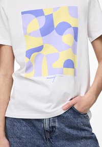 Bílý bavlněný tričko s barevným abstraktním potiskem v modré, žluté a levandulové. Krátké rukávy a volný střih s nezaměnitelným detailem.