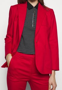 Červený přizpůsobený blejzr se strukturovaným designem, zapínání na jeden knoflík a přední kapsou. V kombinaci s polo tričkem černobíle pruhovaným.