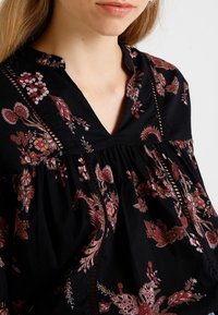 Femme portant un chemisier noir avec des motifs floraux rouges, roses et beiges et des insertions décoratives en dentelle verticale.