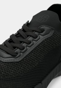 Μαύρο αθλητικό παπούτσι με υφασμάτινο επάνω μέρος με υφή, διαμορφωμένα διακοσμητικά στοιχεία και στρογγυλά κορδόνια. Η εξωτερική σόλα έχει τμηματικό σχέδιο για πρόσφυση.