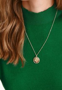 Guldhänge med ett blommönster, hängande på ett fint kedja, buren över en grön ribbad tröja.