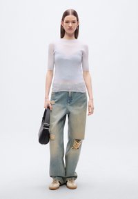 Ανοιχτό γκρι ριμπ κοντομάνικη μπλούζα, φαρδιά ξεθωριασμένα μπλε τζιν με σκισίματα, μπεζ αθλητικά παπούτσια και μαύρη τσάντα ώμου. Απλό φόντο.