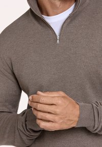 Brauner, strukturierter Strickpullover mit hohem Kragen und Reißverschluss, der gerippte Bündchen hat. Die Hände des Models liegen zusammen vorne.