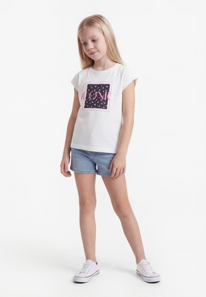 Blond meisje draagt een wit T-shirt met de opdruk "ICONIC", denim shorts en witte sneakers, staat met ontspannen handen voor zich op een witte achtergrond.