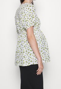Blouse blanche à motifs floraux, ornée de petites fleurs jaunes et de feuilles vertes, avec une taille froncée, des manches courtes bouffantes et une coupe décontractée.