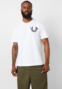 Camiseta de algodón blanca con cuello redondo, mangas cortas y un logo bordado en navy en el lado izquierdo del pecho. Usada con pantalones verde oliva.