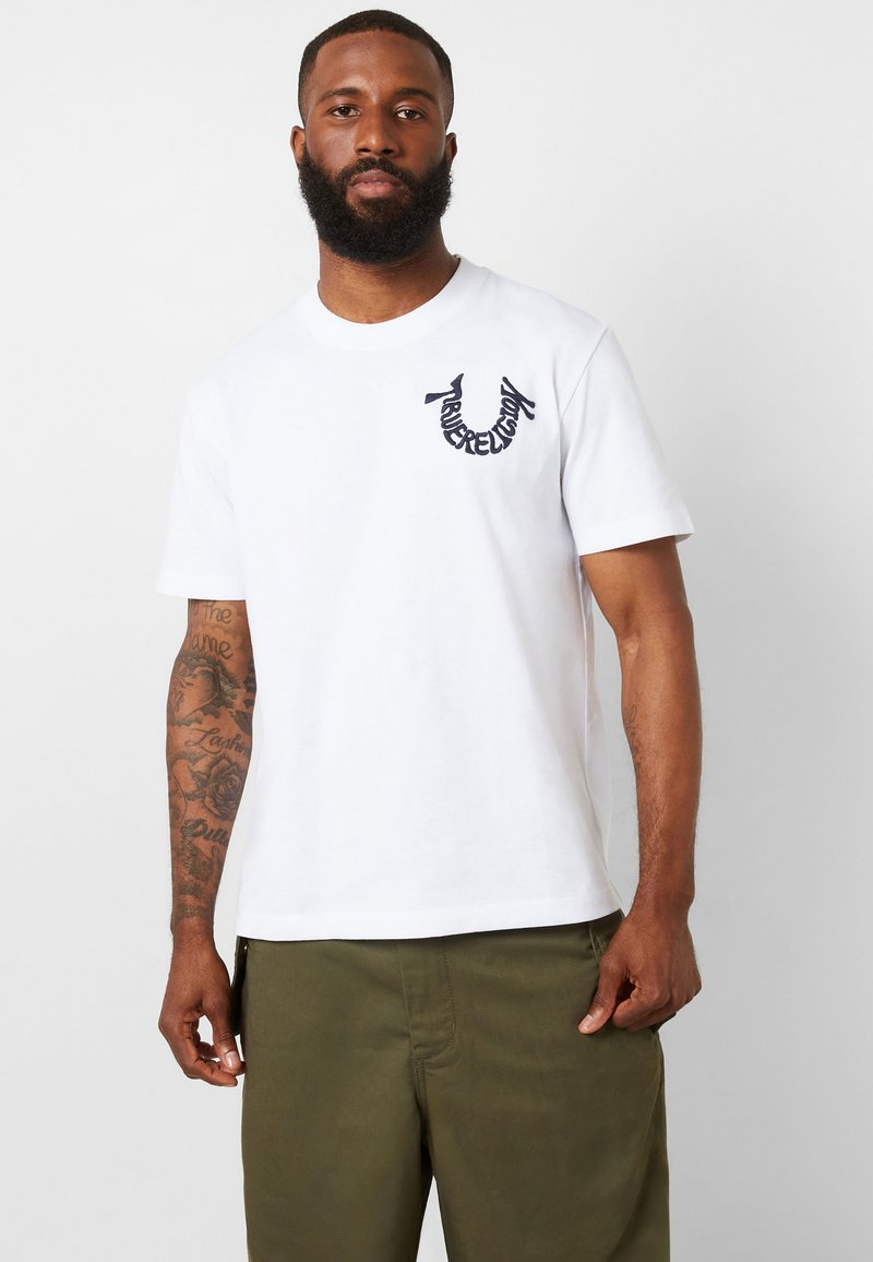 Camiseta de algodón blanca con cuello redondo, mangas cortas y un logo bordado en navy en el lado izquierdo del pecho. Usada con pantalones verde oliva.