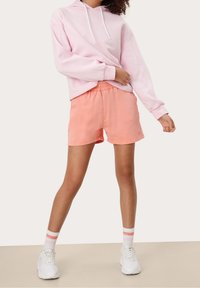 Felpa rosa chiaro con coulisse bianca, abbinata a pantaloni corti pesca e sneakers bianche. L'outfit presenta una vestibilità rilassata e tessuti morbidi.