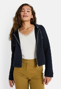 Femme portant une veste matelassée zippée bleu marine, un haut en dentelle blanche et un pantalon couleur moutarde, souriante avec des cheveux ondulés et lâches.