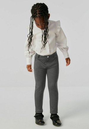 Weißes Hemd mit Rüschendetails an den Schultern, kombiniert mit grauen, eng anliegenden Hosen. Einfarbig und mit glatter Textur, getragen mit schwarzen Schuhen.