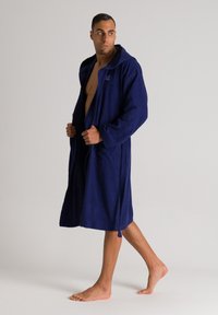 Accappatoio blu navy in tessuto di spugna, con cappuccio, maniche lunghe e due tasche frontali. Il modello è a piedi nudi, mostrando una vestibilità comoda.