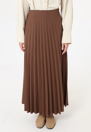 Donna con gonna lunga plissettata marrone a vita alta abbinata a una camicia beige a maniche lunghe e mocassini color cammello.