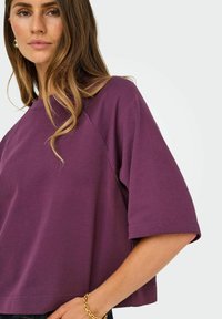 Femme avec de longs cheveux bruns portant un haut violet à manches courtes ample et un bracelet en chaîne dorée, sur un fond clair uni.