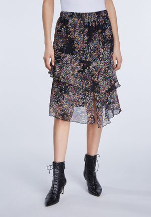 Femme portant une jupe midi superposée noire à fleurs avec des volants et des bottines à lacets noires à bout pointu, debout sur un fond neutre.