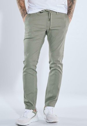 Mann trägt hellolivgrüne Hose mit Kordelzug und weiße Turnschuhe, steht mit den Händen in den Taschen vor einem einfarbigen Hintergrund.