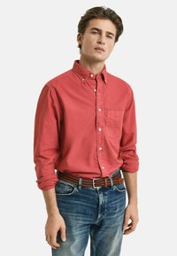 Rotes Button-Down-Hemd mit einer Brusttasche, aus Baumwollstoff, langärmelig mit aufgerollten Ärmeln bis zu den Ellenbogen, getragen mit blauen Jeans und einem braunen Gürtel.