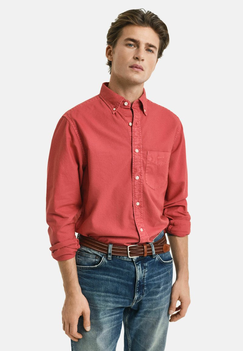 Rotes Button-Down-Hemd mit einer Brusttasche, aus Baumwollstoff, langärmelig mit aufgerollten Ärmeln bis zu den Ellenbogen, getragen mit blauen Jeans und einem braunen Gürtel.