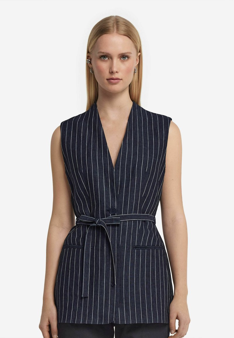 Marinblå ärmlös väst med vita vertikala ränder, V-ringad design, knappstängning och bälte i midjan. Har framfickor och skräddarsydd form.