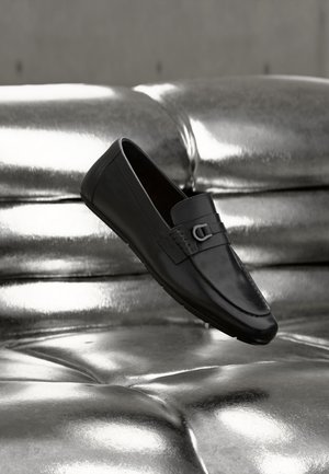Zwarte leren loafer met metalen gespsdetail, geplaatst op een glanzend metalen getufte ondergrond.