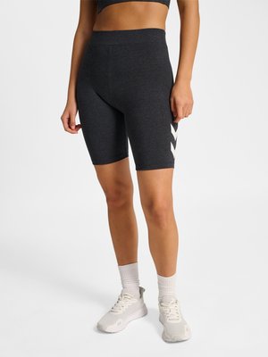 Femme portant un short de sport ajusté gris foncé avec des logos chevrons blancs sur le côté, des chaussettes blanches et des baskets gris clair, debout sur un fond blanc.