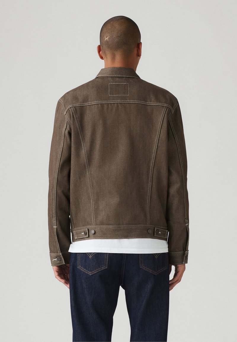 Giacca di jeans marrone con cuciture bianche, design aderente, polsini con bottoni e regolatori laterali. Indossata sopra una camicia bianca e jeans scuri.