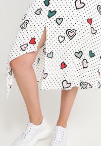 Abito bianco con pois neri e motivi a cuore colorati; spacco laterale; indossato con sneakers bianche. Tessuto leggero con texture morbida.