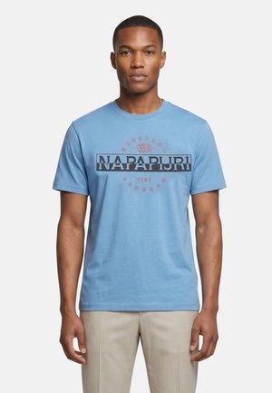 Homme portant un t-shirt Napapijri bleu clair et un pantalon beige, debout sur un fond blanc uni.
