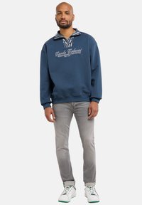 Laivastonsininen collegepaita, jossa kuvioitu kaulus ja valkoinen kirjailtu teksti. Yhdistetty vaaleanharmaisiin farkkuihin ja valkoisiin lenkkareihin, joissa on vihreitä yksityiskohtia.