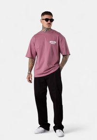 Mann mit Tattoos und Sonnenbrille, der ein lockeres mauvefarbenes T-Shirt, schwarze Hosen und weiße Sneaker trägt, steht vor einem weißen Hintergrund.