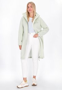 Donna in giacca lunga zip-up verde chiaro, pantaloni bianchi a vita alta, maglietta a righe e sneakers beige, in piedi contro uno sfondo bianco.