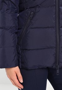 Giacca trapuntata blu navy con tasche anteriori visibili e cerniere angolate, indossata da una persona con la mano appoggiata accanto all'anca.