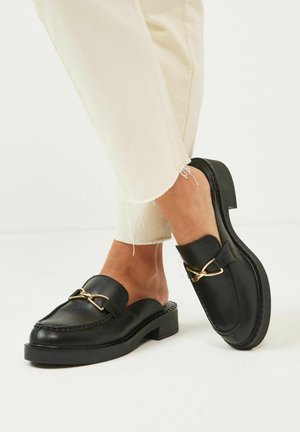 Mocassins noirs en cuir à enfiler avec une boucle dorée portés avec un pantalon court crème sur un fond clair.