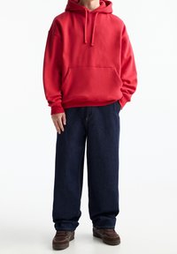 Czerwona bluza z kapturem i kieszenią z przodu, w połączeniu z luźnymi, ciemnoniebieskimi dżinsami. Stylizację uzupełniają brązowe sneakersy.