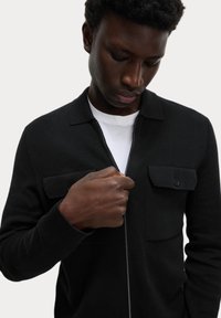 Sort zip-up cardigan med to brystlommer. Har en krave og ribbede manchetter, lavet af et glat, struktureret stof.
